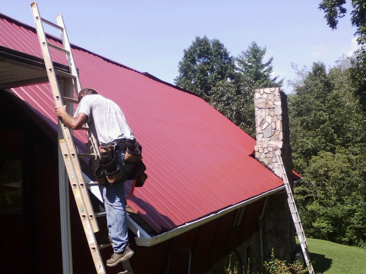 Roofer on a ladder beside a red metal roof and stone chimney — Soffit & Fascia Repair by Roofing Brothers in Mansfield